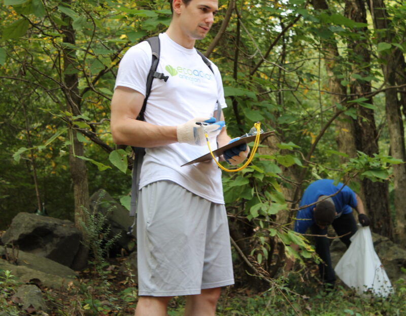 square image with volunteer in foreground wearing a white EcoAction Arlington t-shirt and work gloves holding a clipboard and pen, in the foreground is a forested area with green fauna, trees and boulders, in the bottom right a volunteer in a blue t-shirt holding a white trash bag is bending down to pick up trash