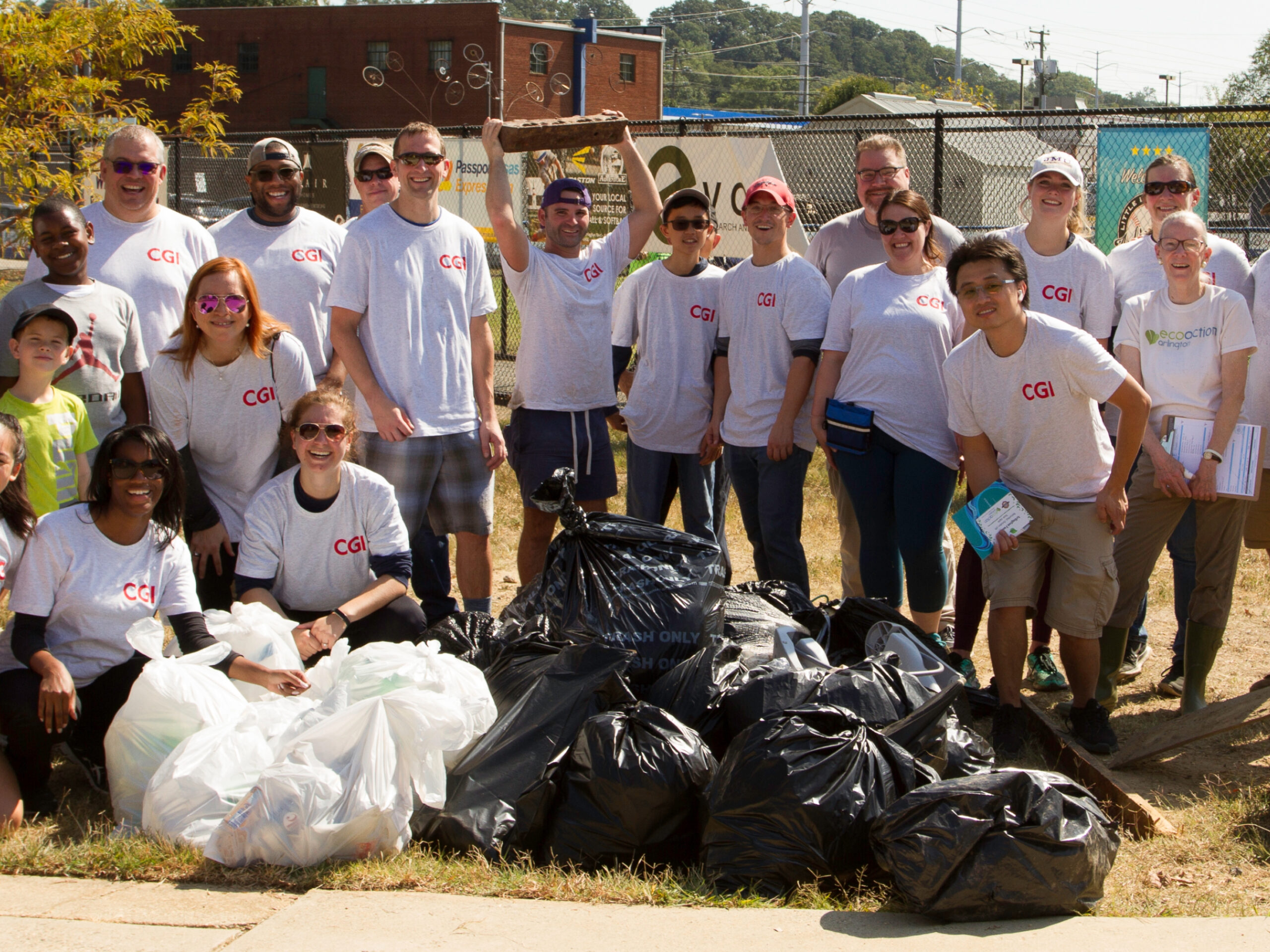 Group of volunteers of different ages in white t-shirts standing in front of several black trash bags and large white recycling bags outside in a local park