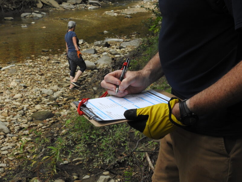 image of a torso with hands and arms holding a clipboard and noting trash collected with a pen, in the background is a stream and a volunteer picking up trash