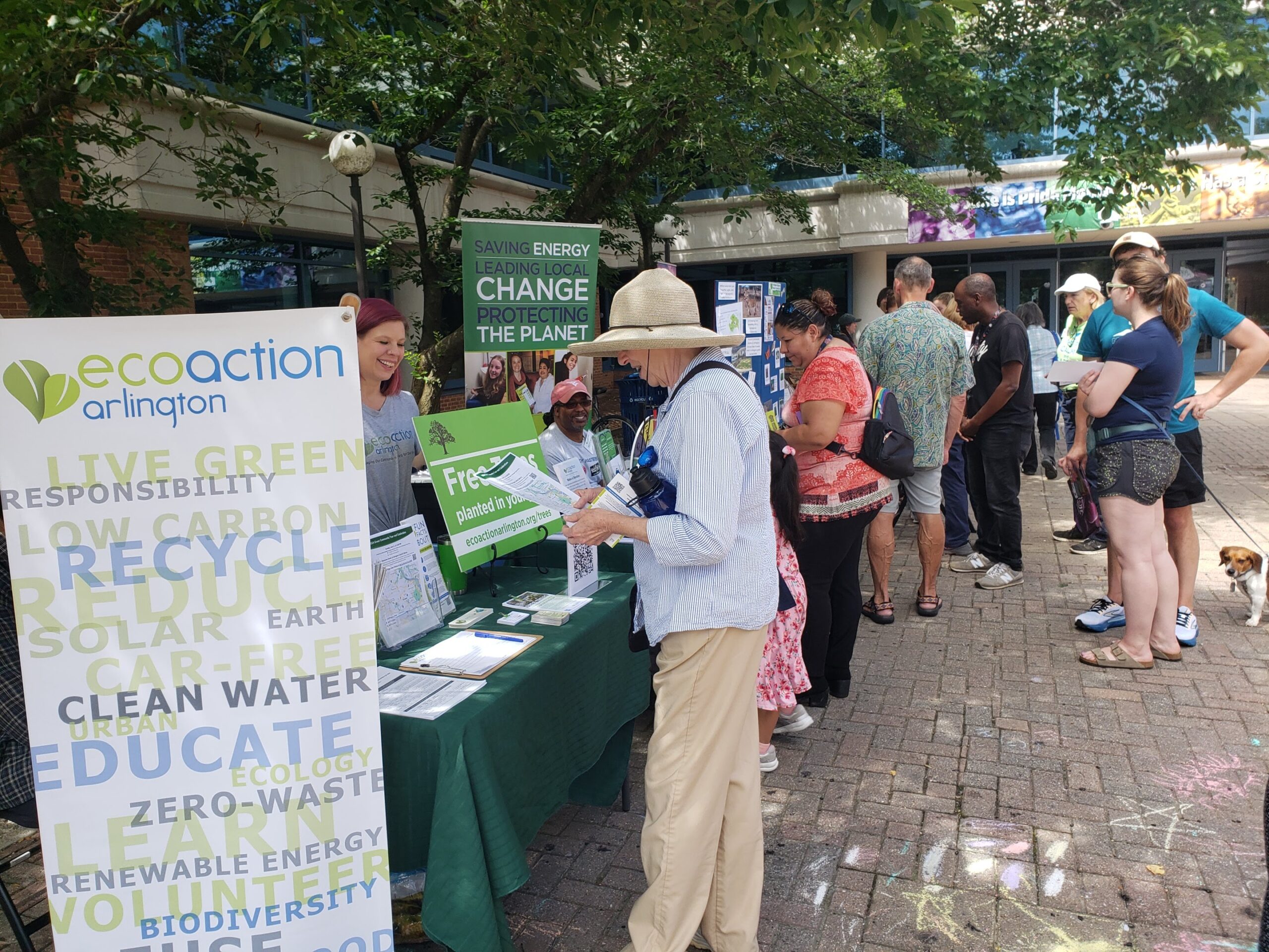 woman in white tshirt, brimmed hat and beige pants stopping in front of the EcoAction Arlington table on display outside of the Central Library in Arlington. Kara Simon greets her from behind the table