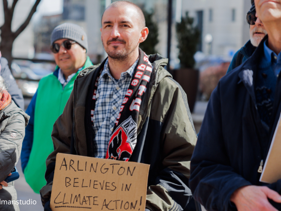 six attendees of the rally on 2/22/205 gathered to hear speakers. A man in the front and center in blue and with plaid shirt, scarf and black bomber jacket holds a sign that reads: Arlington believes in climate action!