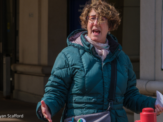 Close up of Julie Rosenberg, Faith Alliance for Climate Solutions, in a teal parka with arms held out speaking to the crowd. Photo credit: Bryan Scafford.