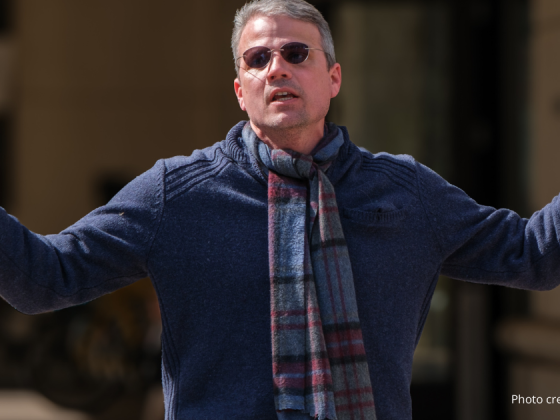 Kip Malinowsky, teacher at Thomas Jefferson Middle School, in a navy sweater and blue tartan scarf and arms held up wide addressing the crowd at the 2/22/2025 rally. photo credit: Bryan Scafford.