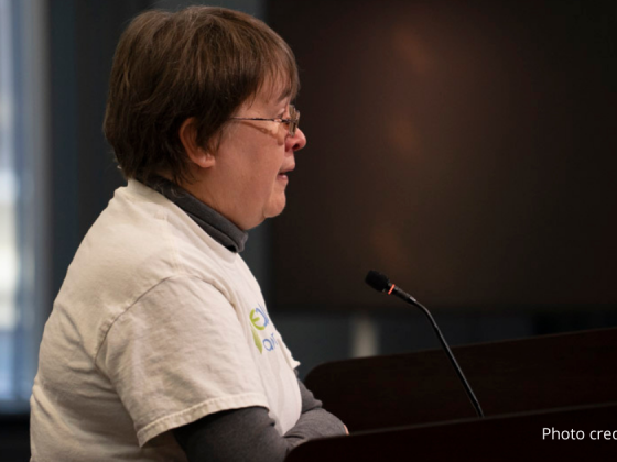 Joan McIntyre, EcoAction Arlington board member, wearing a white t-shirt with the ecoaction arlington logo, speaking at the podium in front of Arlington County Board on 2/22/2025. Photo credit: Molly Bolan.