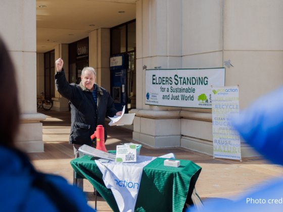 Jim Lyon, EcoAction Arlington board member, holding up right fist and holding papers in left hand speaking in front of the crowd. There is a table with the Ecoaction Arlington branded green and white tablecloth, and red megaphone on table. In the background is the Bozman Government Center building, Elders Standing for a Sustainable and Just world banner sign, and an EcoAction Arlington sign. in the foreground are closeups over the shoulders of two rally-goers.