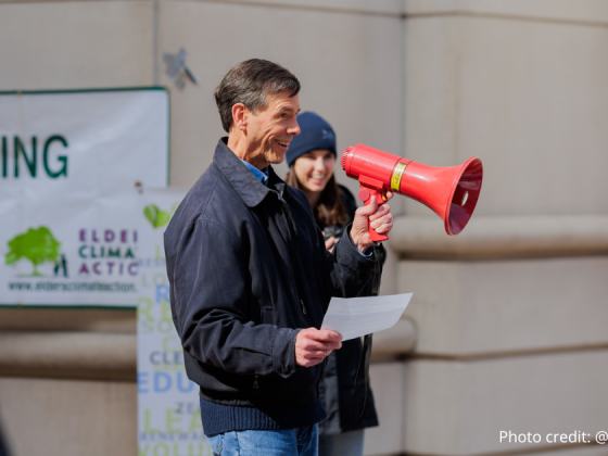 John Bloom of Sierra Club Potomac River Group holding a red megaphone and an piece of paper, smiling and addressing the crowd. Molly Bolan is seen in the background. Photo credit: @elmanstudio.